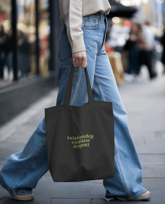 Millenial holding a black tote bag with text saying "relationship escalator dropout" on a city street