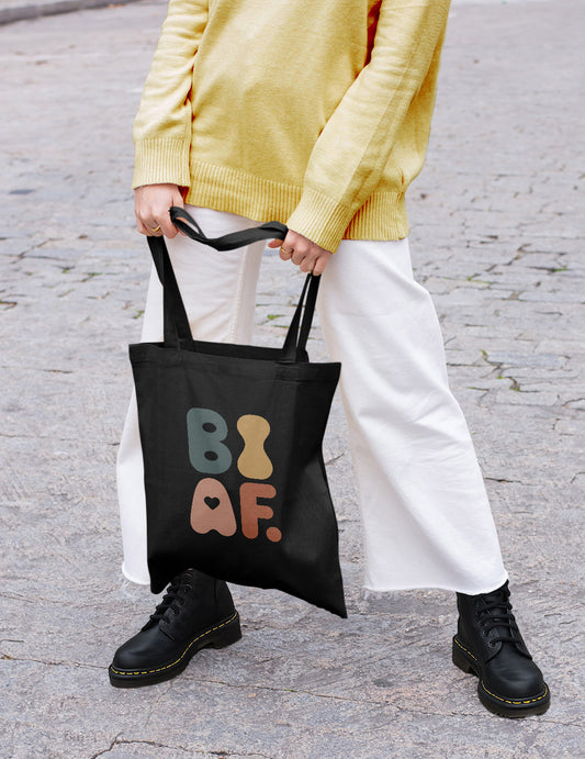 A person in a yellow sweater, loose jeans and Doc Martens holds a colorful LGBTQ+ tote bag. The setting is a cobblestone street.