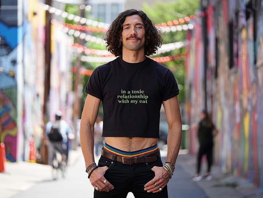 Male model wearing black crop top with sarcastic "in a toxic relationship with my cat" green quote. Colorful street setting.