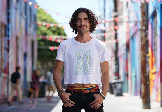 Male model wears a white neurodivergent affirmation crop top with smileys design while standing in a colorful city alley. He has curly hair and a mustache.