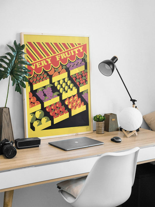 Vintage-inspired queer humor poster with vibrant fruit stand, displayed on a desk in a sunlit office with a laptop, notebook, and coffee, creating a modern, creative vibe.