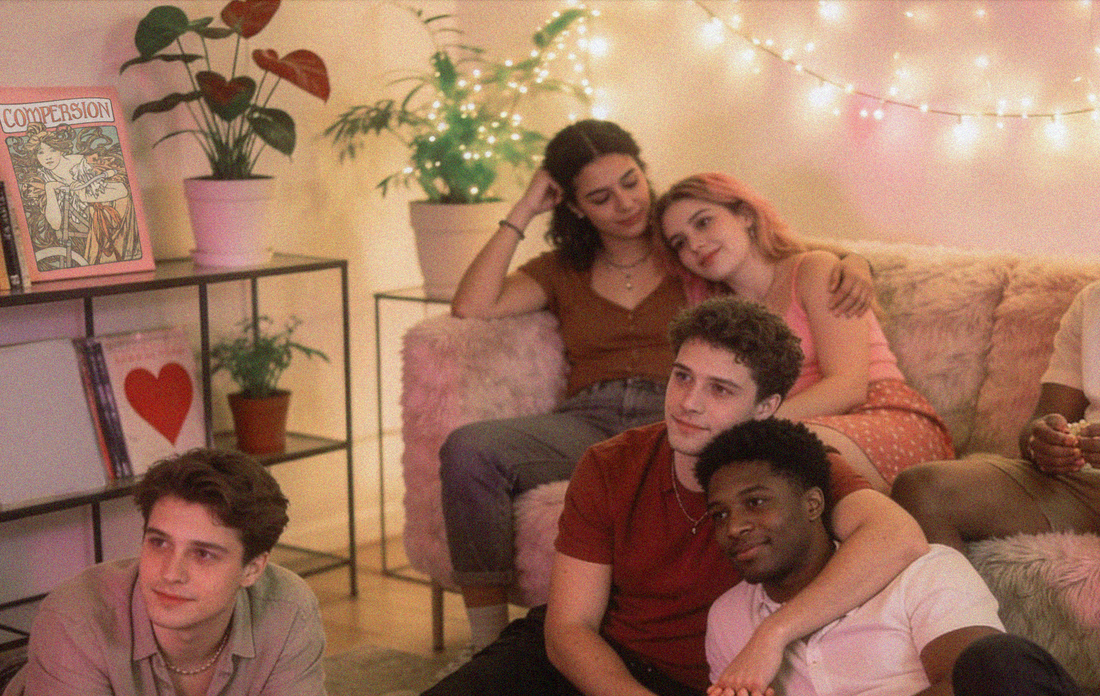 A cozy room with soft lighting and string lights, five queer polyamorous people are gathered. Two are cuddling on a sofa, while three sits contently on the floor.