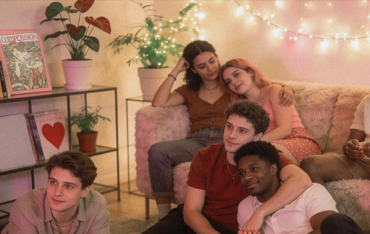 A cozy room with soft lighting and string lights, five queer polyamorous people are gathered. Two are cuddling on a sofa, while three sits contently on the floor.