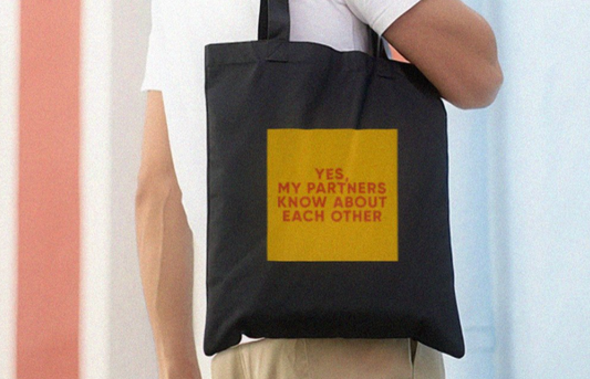 A polyamorous person holds a black tote bag with a yellow square featuring red text reading, "Yes, my partners know about each other." The scene conveys joy and openness.