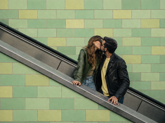 A couple kisses on an escalator against a green tiled wall. They wear casual jackets, conveying love and spontaneity in an urban setting.