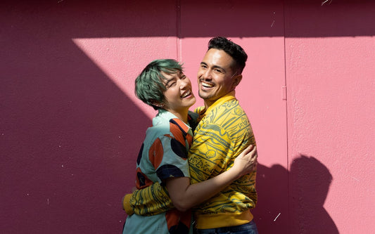 A joyful, non-normative couple embraces, wearing colorful outfits against a vibrant pink wall. They are smiling and laughing, radiating happiness and affection.