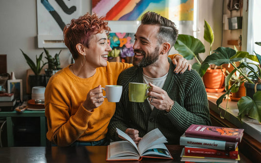A smiling ENM couple sits at a table, holding mugs with their arms around each other. Open books and vibrant plants create a cozy, warm ambiance.