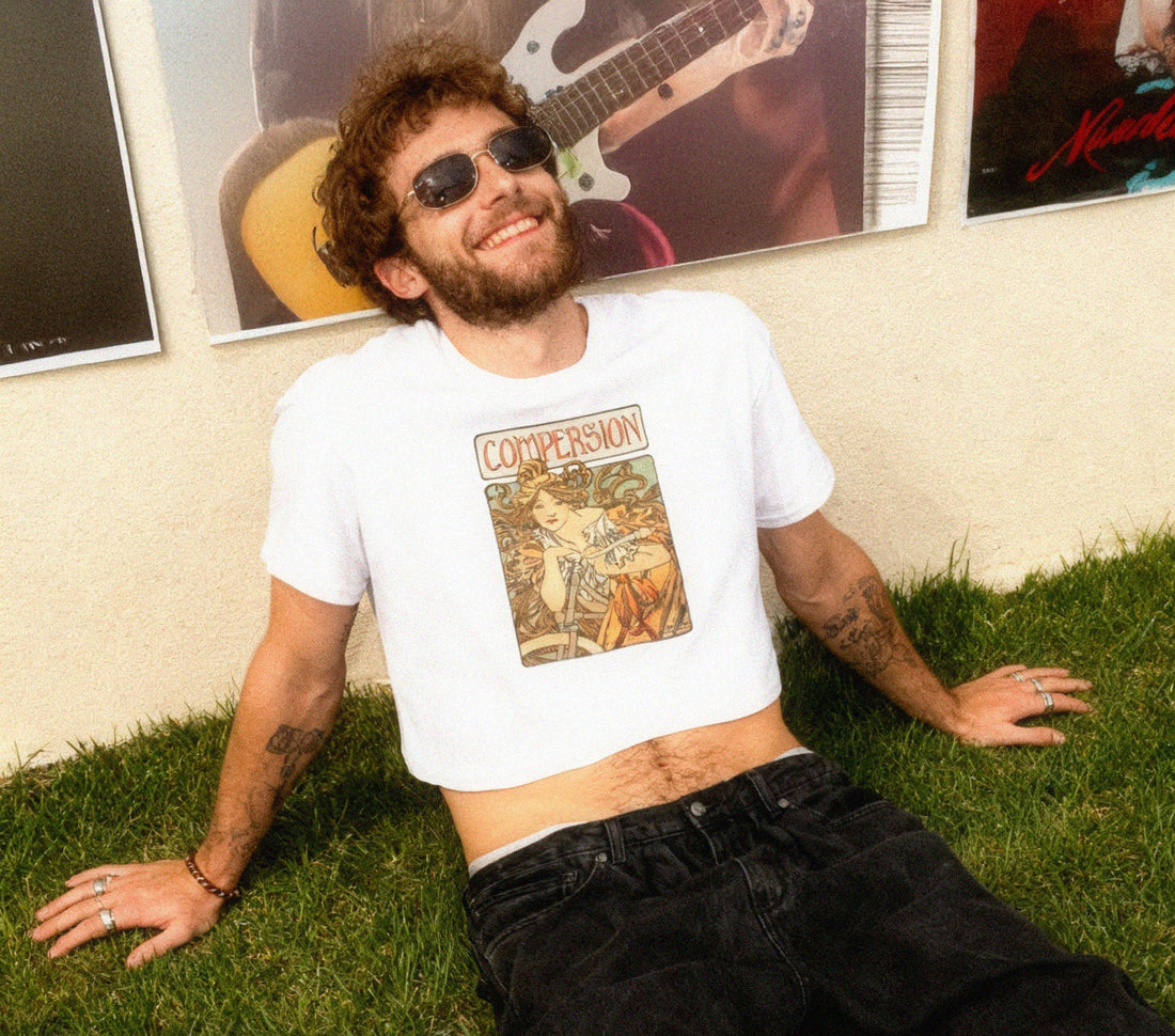 Man confidently wearing a crop top, smiling and posing sitting on the grass with posters behind him.