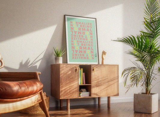 A cozy room with a wooden cabinet holding books, a plant, and a framed print saying "Normalize Not Being Normal" for neurospicy affirmation. Soft light and a potted palm add warmth.