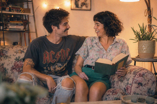A joyful, non-normative couple embraces, wearing colorful outfits in a cozy living room. They are smiling and laughing, radiating happiness and affection.