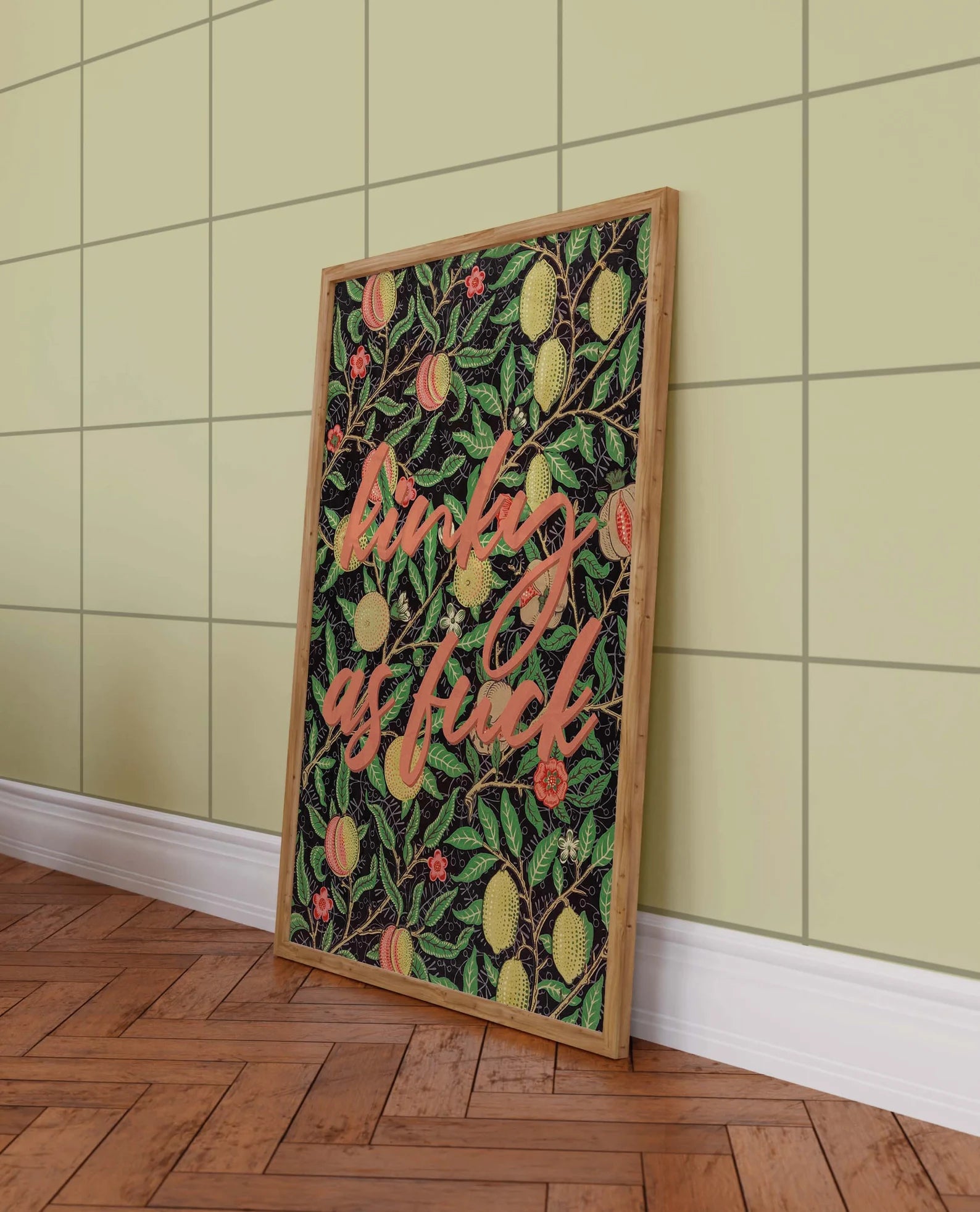 Framed vibrant poster with text printed in bold colors for pleasure-centered living leans against a green wall on a wooden floor.