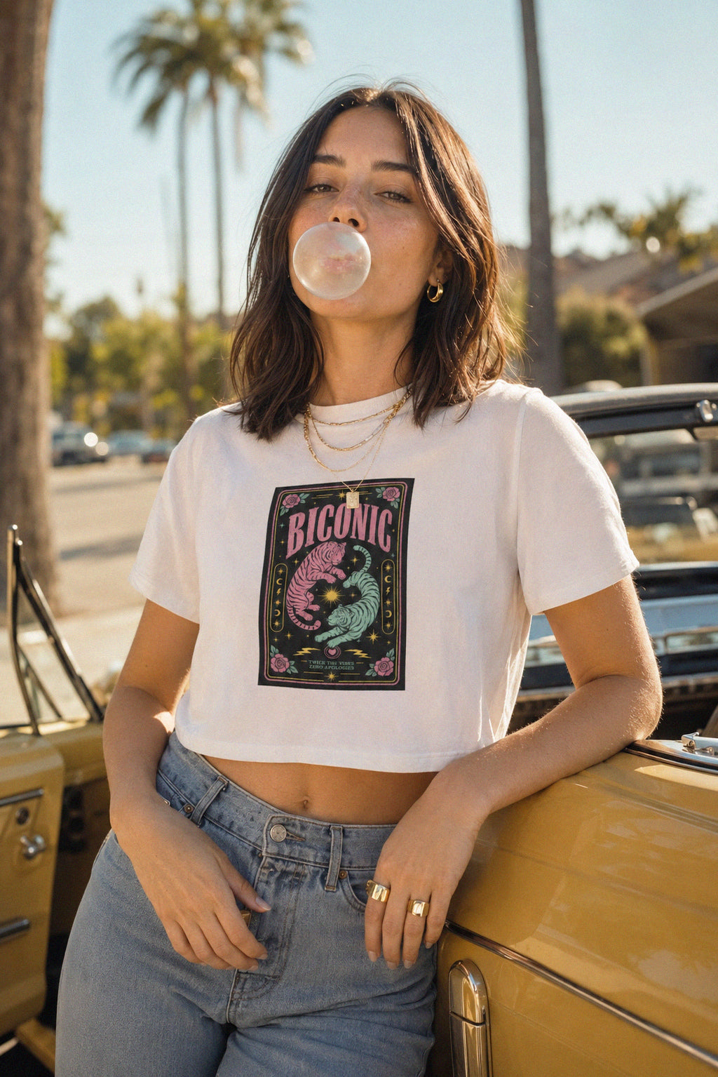 A woman wearing a white crop top t-shirt with a colorful mermaid graphic that reads 'BICONIC' and blue jeans, leaning against a yellow vintage car outdoors with palm trees in the background.