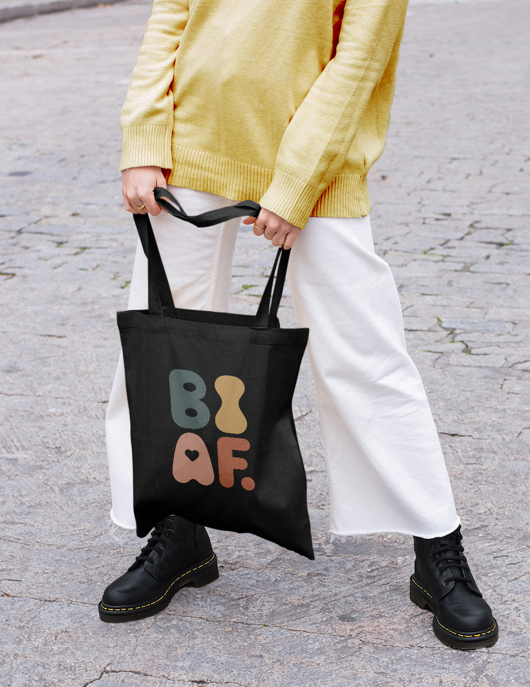 A person in a yellow sweater, loose jeans and Doc Martens holds a colorful LGBTQ+ tote bag. The setting is a cobblestone street.