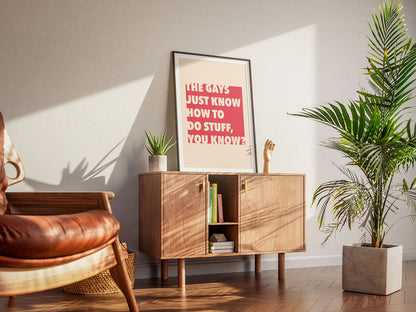 A cool living room with a framed colorful red LGBTQ+ printable poster with iconic pop culture quote. A wooden armchair, a cabinet and houseplants create a warm, inviting atmosphere.