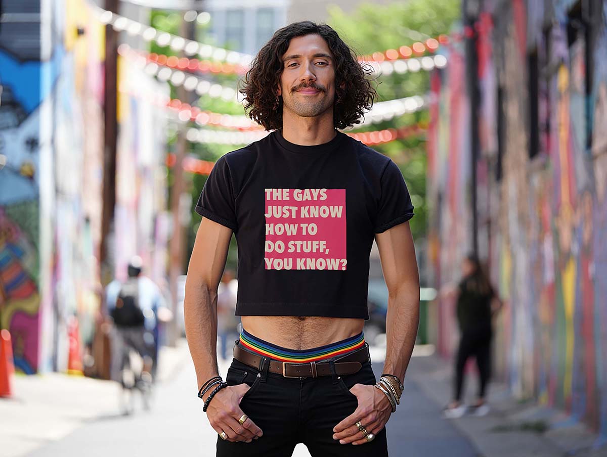 Male model wears a queer White Lotus quote crop top while standing in a colorful city alley. He has curly hair and a mustache.