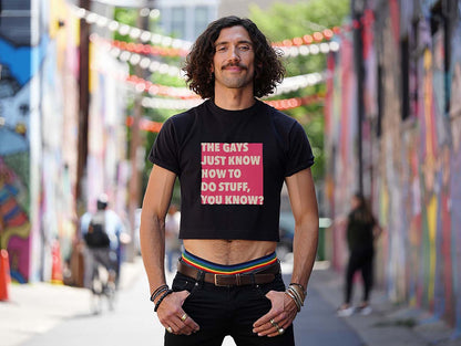 Male model wears a queer White Lotus quote crop top while standing in a colorful city alley. He has curly hair and a mustache.
