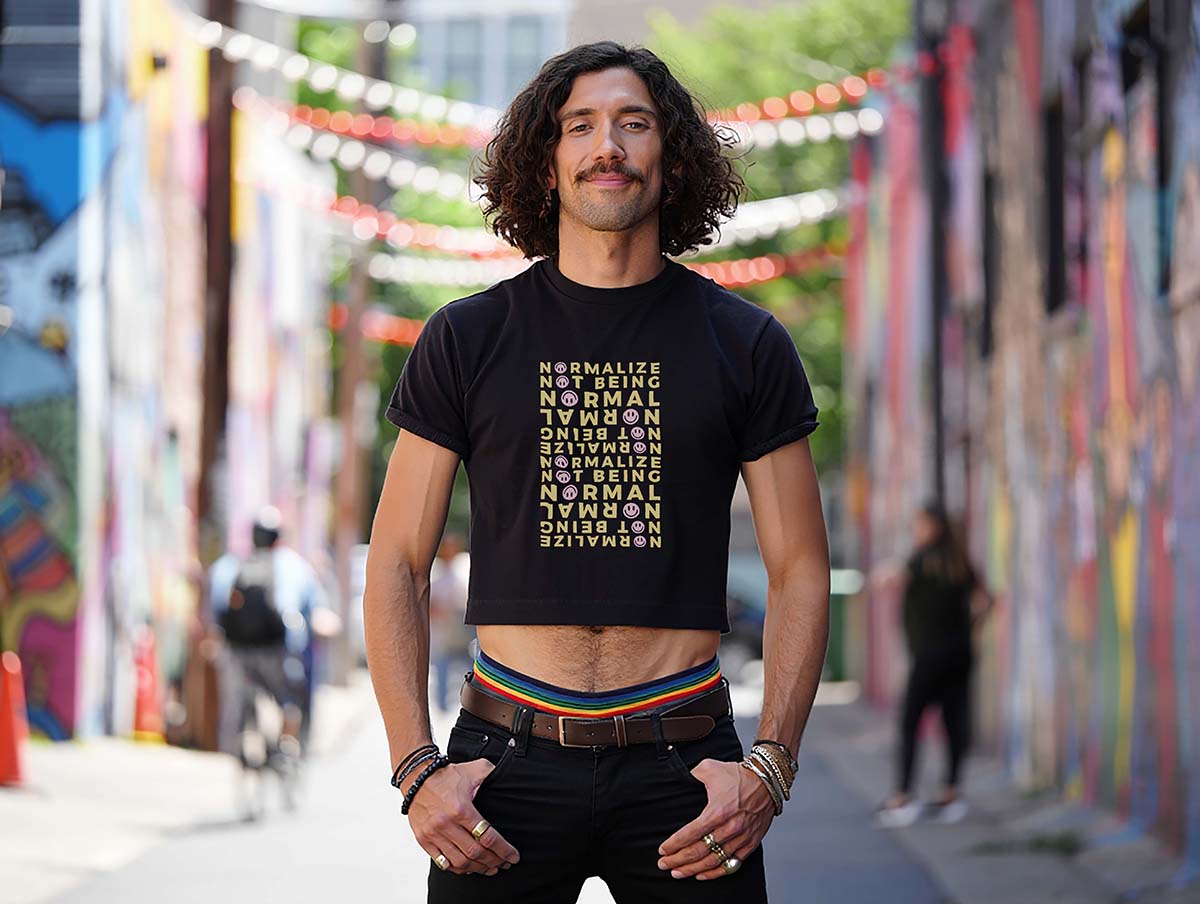 Male model wears a bold neurodivergent affirmation crop top with smileys while standing in a colorful city alley. He has curly hair and a mustache.