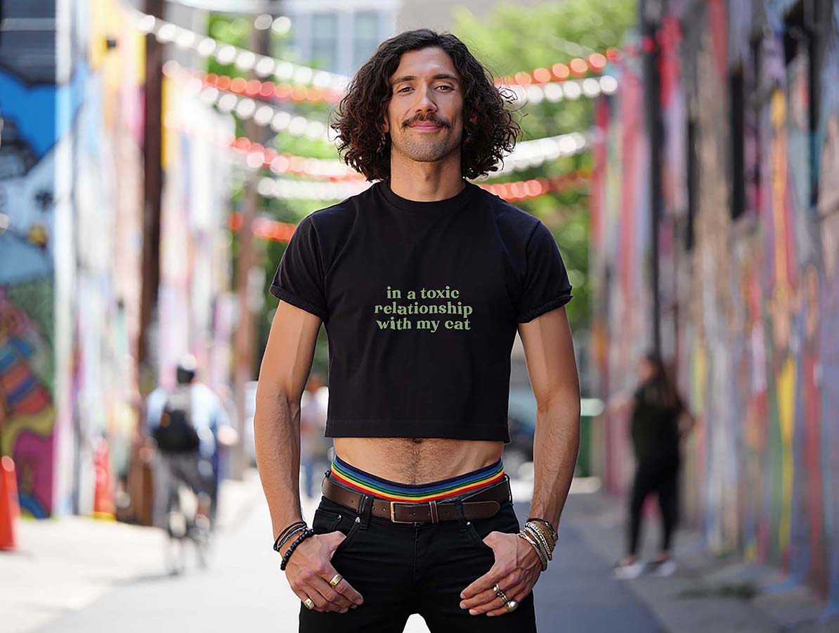 Male model wearing black crop top with sarcastic "in a toxic relationship with my cat" green quote. Colorful street setting.