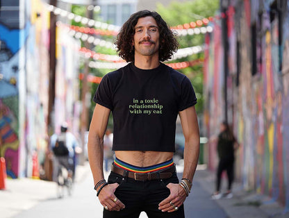 Male model wearing black crop top with sarcastic "in a toxic relationship with my cat" green quote. Colorful street setting.