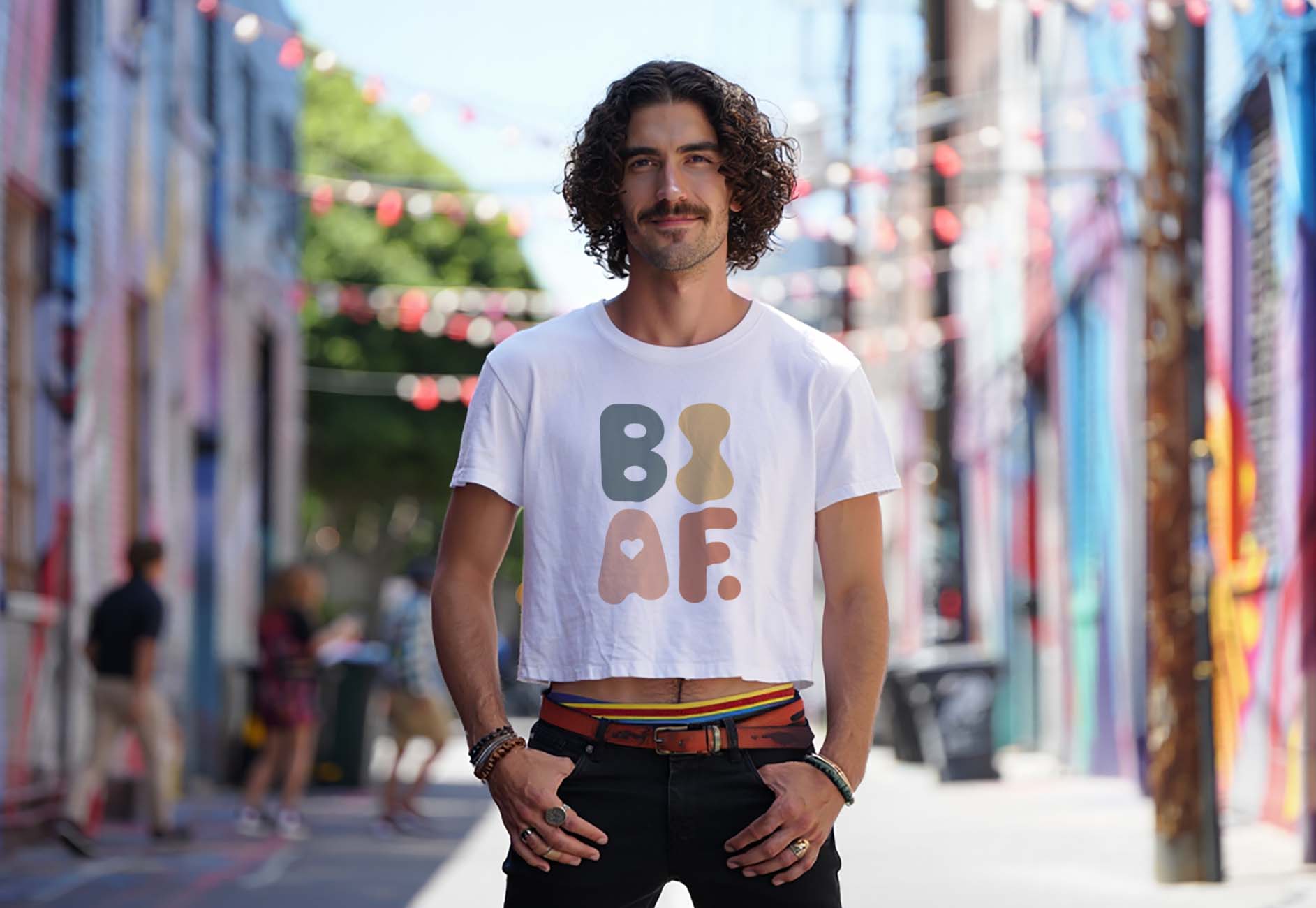 A man with curly hair and a mustache stands in a colorful, urban alley. He wears a white crop top with bold BI AF letters with heart design