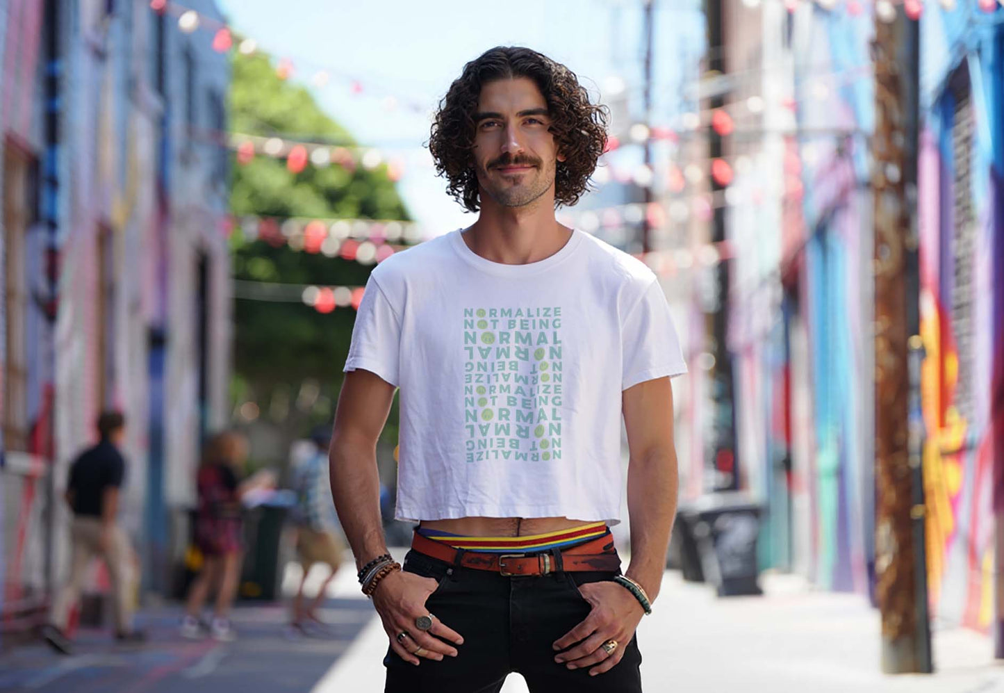 Male model wears a white neurodivergent affirmation crop top with smileys design while standing in a colorful city alley. He has curly hair and a mustache.