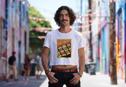 Smiling male model with curly hair and mustache in street alley wearing a white crop top with colorful "Very fruity" vintage design. For queer visibility with edge.