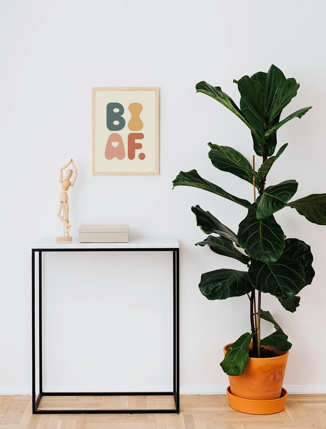 A framed queer poster reading "BI AF" in bold, joyful colours artwork above a minimalist interior with a tall green plant in an orange pot next to a small black table.