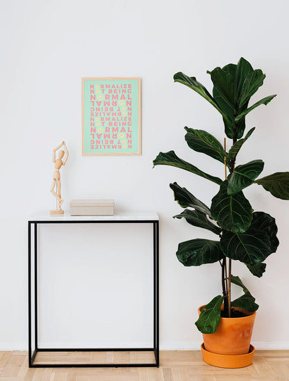 A framed modern Neurodivergent Poster with green tones above a minimalist interior with a tall green plant in an orange pot next to a small black table.