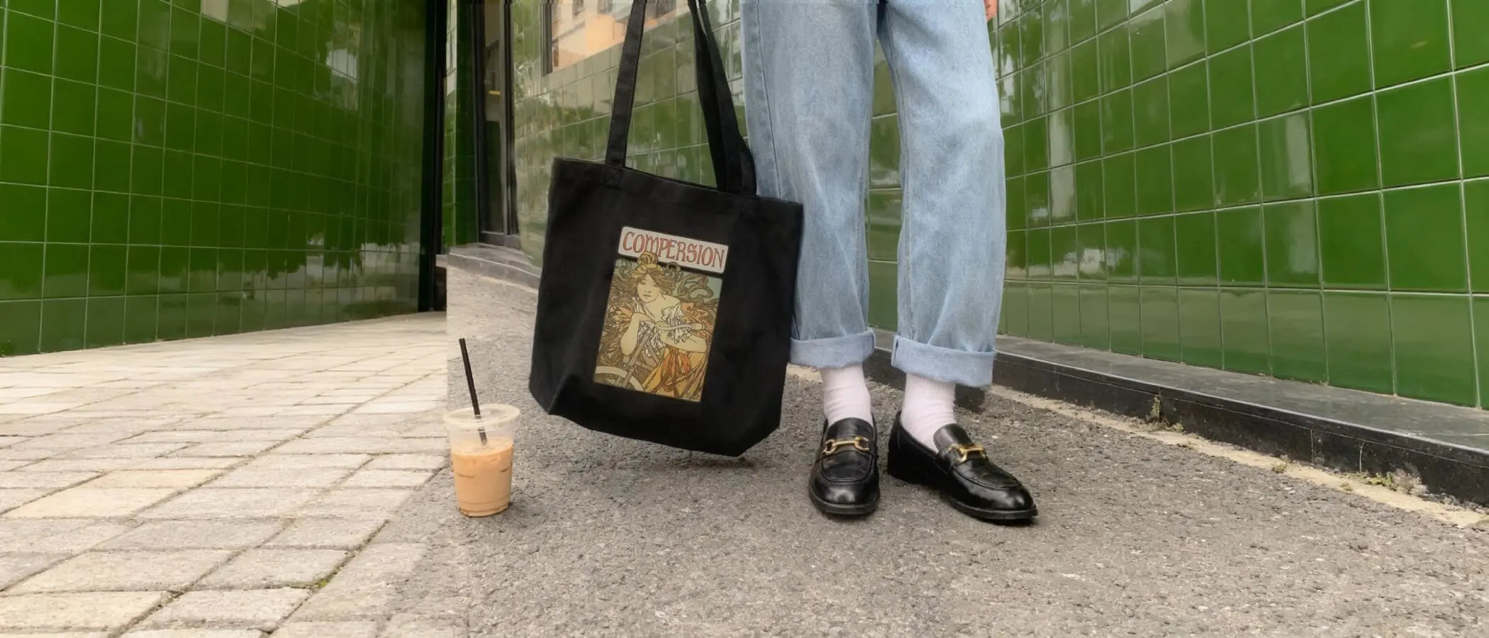 Fashionable person in cuffed jeans stands against a green tiled wall. Black loafers and white socks are visible. They hold a black tote with word "COMPERSION" for ENM affirmation and visibility.
