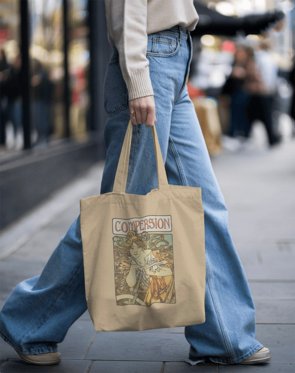 A person walks down a city street carrying a stylish Compersion beige tote bag showcasing ENM relationship. They wear a casual style sweater and loose jeans.